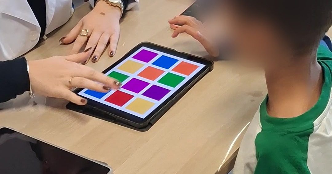 Mãos da terapeuta e de uma criança junto a um tablet com quadrados coloridos, durante uma sessão